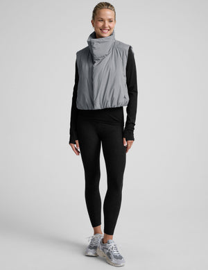 model wearing a gray puffer vest over a black long sleeve and leggings on a light gray background. 