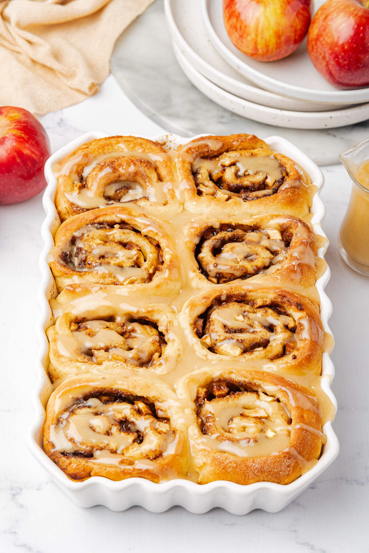 Overhead view of a full dish of warm cinnamon rolls, topped with a shiny glaze, with apples and plates nearby.