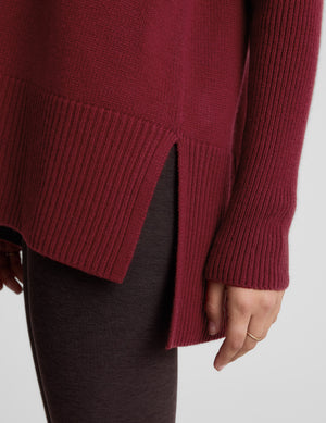 Woman wearing a red v-neck long length sweater and brown leggings against a plain background. 