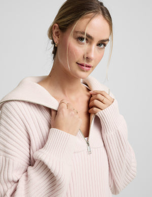 Woman wearing a light pink quarter zip sweater and white fleece pants against a plain background. 